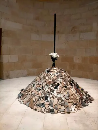 O milladoiro da memoria. Un c&uacute;mulo de pedras coroado por flores ocupa o antigo pan&oacute;ptico do MARCO na exposici&oacute;n Hai un lugar, de Alberto Ardi, como s&iacute;mbolo do punto onde unha vida quedou truncada e, ao mesmo tempo, como ofrenda colectiva &aacute; memoria das v&iacute;timas.