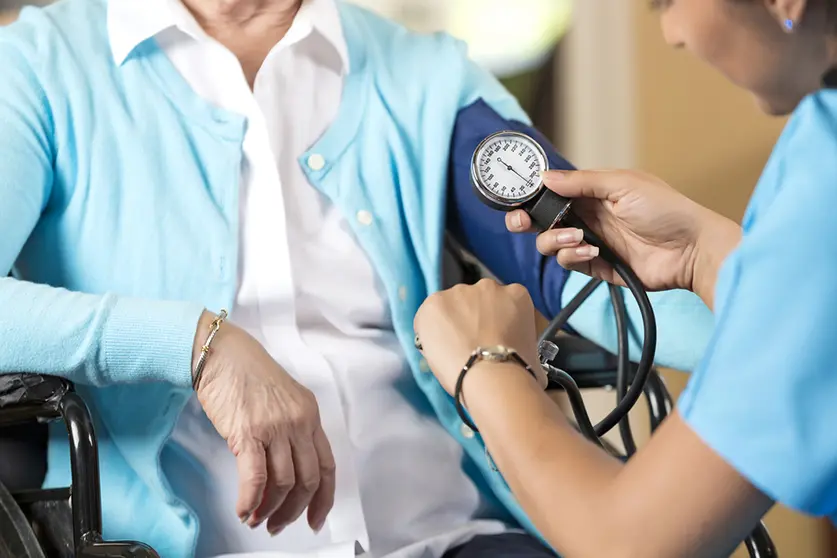 Close up of hands while nurse takes blood pressure of the woman sitting in the wheelchair in front of hair. Senior caucasian woman and a young hispanic woman. They are indoors, sitting down. The nurse is looking at her watch as she is working. The senior woman is wearing a white shirt with a light blue cardigan on top of it, the nurses scrubs are nearly the same color as the womans cardigan.