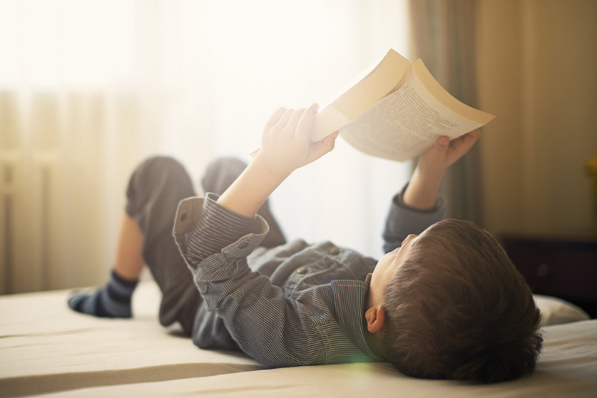 Little boy lying in bed and reading a book. The boy is aged 6. Sunny day.
