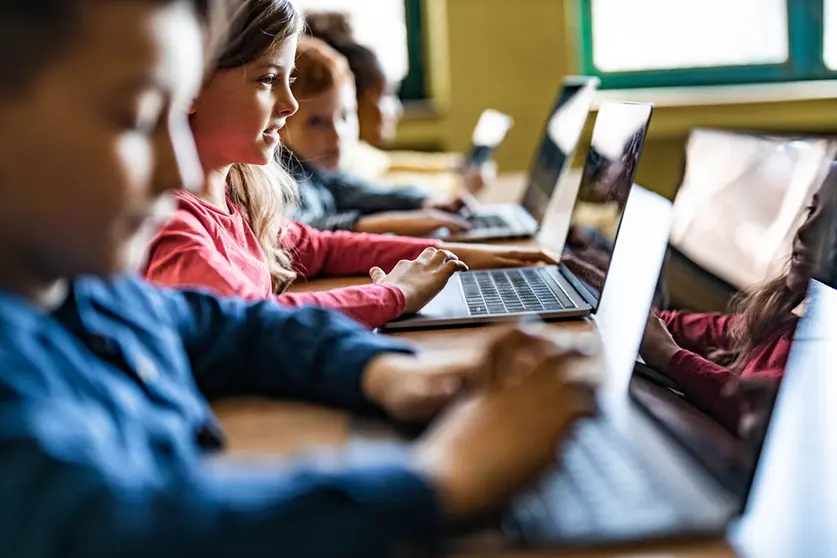 Happy schoolgirl and her classmates e-learning over laptops during a class in the classroom.