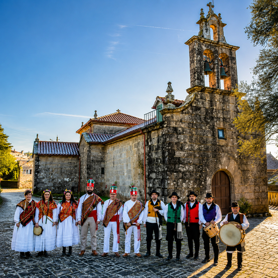 Integrantes da Asociaci&oacute;n Folcl&oacute;rica Pedra da Garza diante da igrexa parroquial de San Bartolomeu de Fozara, s&iacute;mbolo da uni&oacute;n entre patrimonio, m&uacute;sica tradicional e vida veci&ntilde;al.