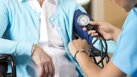 Close up of hands while nurse takes blood pressure of the woman sitting in the wheelchair in front of hair. Senior caucasian woman and a young hispanic woman. They are indoors, sitting down. The nurse is looking at her watch as she is working. The senior woman is wearing a white shirt with a light blue cardigan on top of it, the nurses scrubs are nearly the same color as the womans cardigan.
