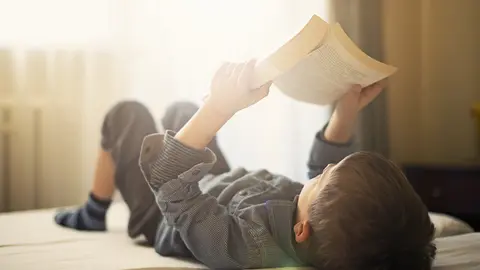 Little boy lying in bed and reading a book. The boy is aged 6. Sunny day.