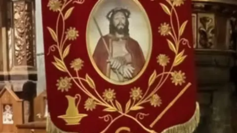 Estandarte de la Hermandad del Cristo Resucitado para la procesi&oacute;n de la Santa Cena, con fondo rojo y simbolog&iacute;a de la Pasi&oacute;n como la corona de espinas y la ca&ntilde;a del Ecce Homo.