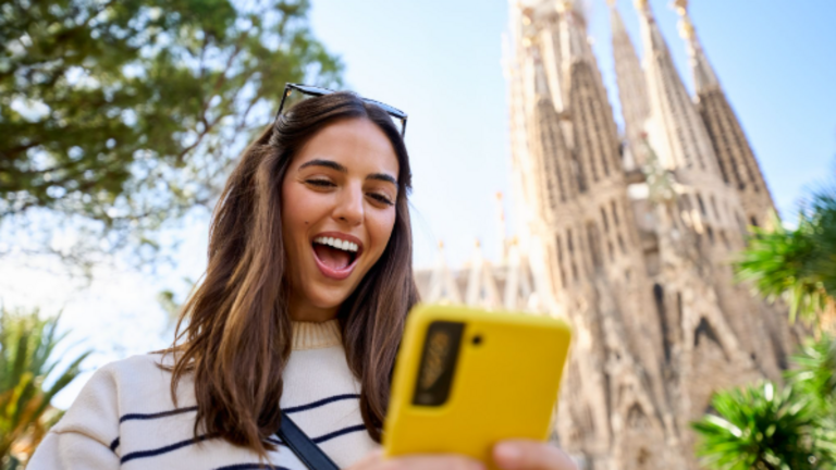 Una joven consulta su tel&eacute;fono m&oacute;vil frente a la Sagrada Familia de Barcelona, una de las ciudades espa&ntilde;olas que concentra un creciente n&uacute;mero de n&oacute;madas digitales en 2026.