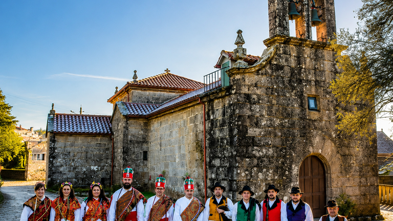 Integrantes da Asociaci&oacute;n Folcl&oacute;rica Pedra da Garza diante da igrexa parroquial de San Bartolomeu de Fozara, s&iacute;mbolo da uni&oacute;n entre patrimonio, m&uacute;sica tradicional e vida veci&ntilde;al.