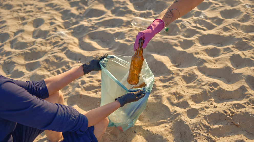 Una persona recoge una botella de pl&aacute;stico de la arena, un gesto sencillo que contribuye a reducir los residuos y proteger los ecosistemas costeros.