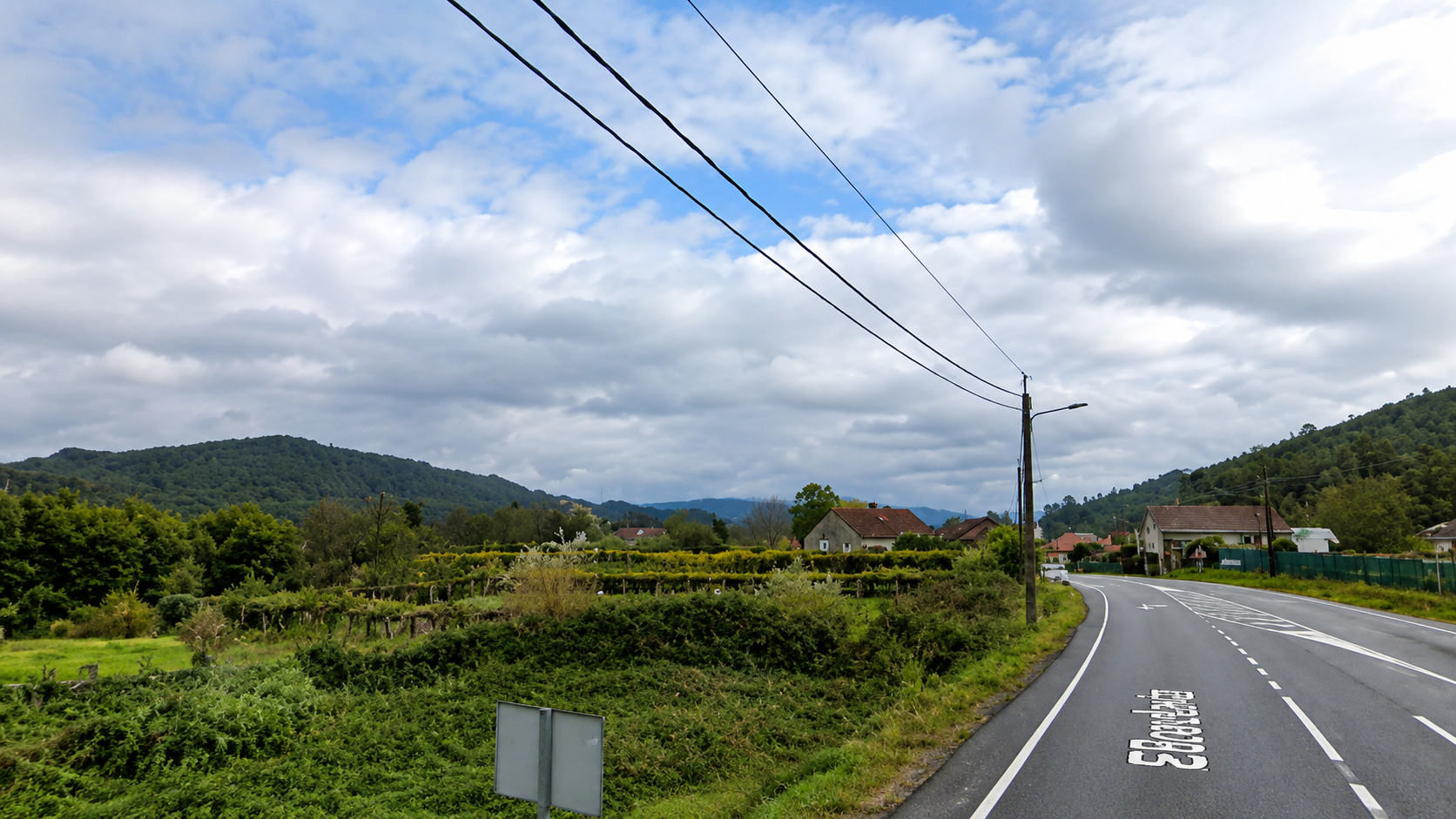 Vista da contorna rural de Pías, coa estrada atravesando unha paisaxe marcada polos montes, as leiras e o ceo cambiante.